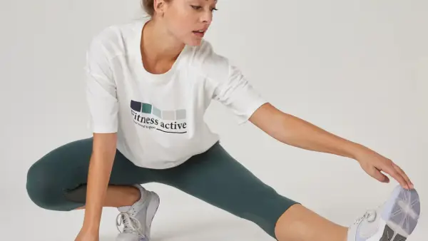 Kobieta w białej koszulce z napisem "Fitness active" wykonuje rozciąganie po treningu siłowym.