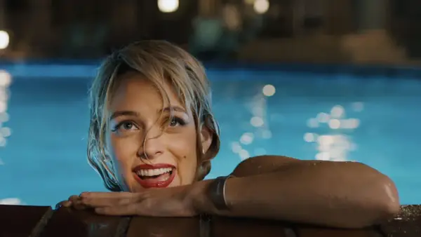 Una donna sorride dall'acqua di una piscina, con luci sfocate sullo sfondo. Una scena che ricorda "Under the Silver Lake".