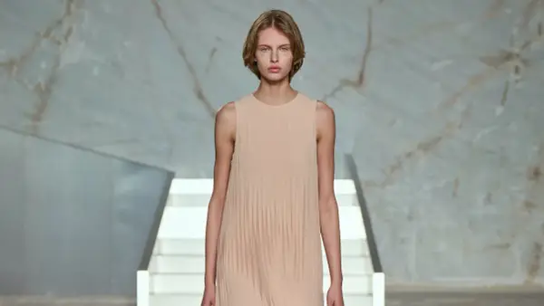 Modelo con vestido plisado color melocotón, en un escenario minimalista con escaleras blancas y fondo de mármol.
