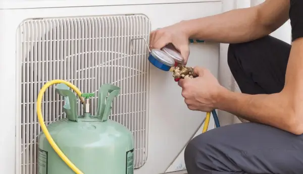 Técnico revisa manómetro para detectar fuga aire acondicionado. Cilindro de gas refrigerante y unidad exterior visibles.