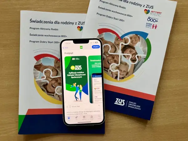 Aplikacja mZUS na telefonie pokazuje świadczenie wychowawcze 800+ dla rodziny. Ulotki ZUS informują o programach dla rodziców.