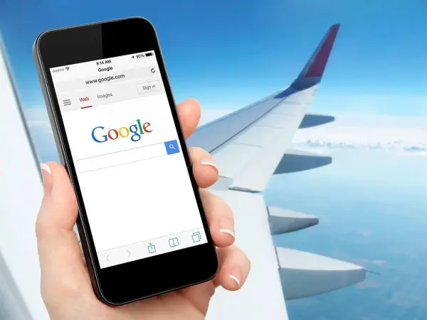 Dłoń trzyma smartfon z otwartą wyszukiwarką Google, na tle skrzydła samolotu i nieba. Czy w samolocie można mieć internet? Tak, można!
