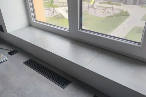 Jasny parapet z płytek wewnątrz, z widokiem na plac zabaw. Obok laptop i kratka wentylacyjna.