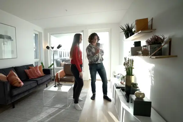 Två personer diskuterar olika inredningsstilar i ett ljust vardagsrum med modern möblering, en mörk soffa och hyllor med växter.