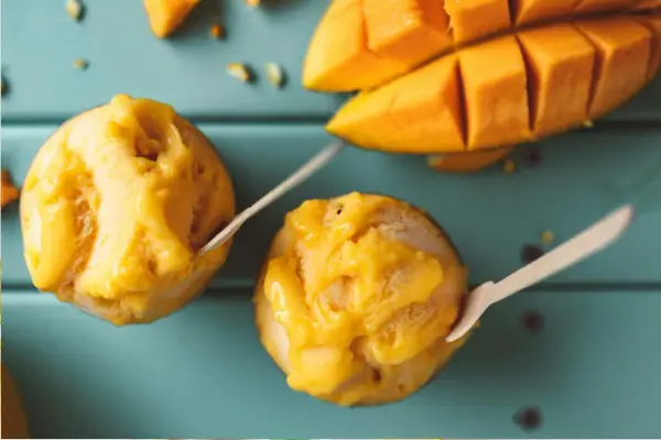 Jak zrobić sorbet? Dwa pucharki z mango sorbetem i kawałki świeżego mango na turkusowym tle.