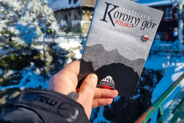 Dłoń trzyma książeczkę "Korona Gór Polski" na tle zimowego krajobrazu.