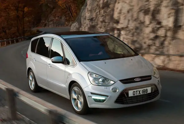 Biały Ford S-Max 2.0 TDCI pokonuje zakręt na malowniczej drodze wśród jesiennych drzew i skał.