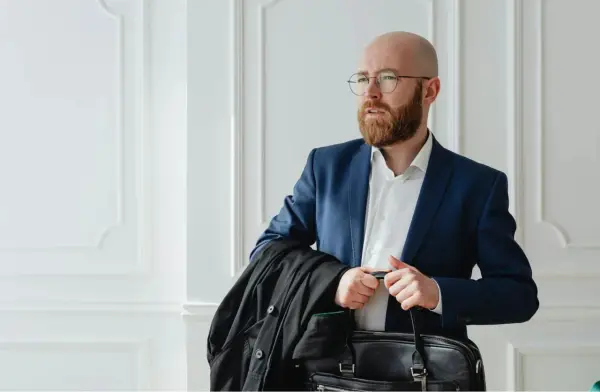 Un homme crane rasé, barbe rousse, lunettes, en costume bleu, porte une veste noire et une mallette.