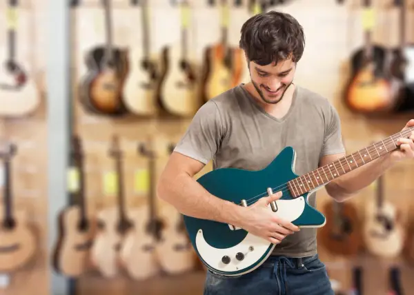 Uśmiechnięty mężczyzna testuje gitarę elektryczną w sklepie muzycznym. W tle widać wiele innych instrumentów.