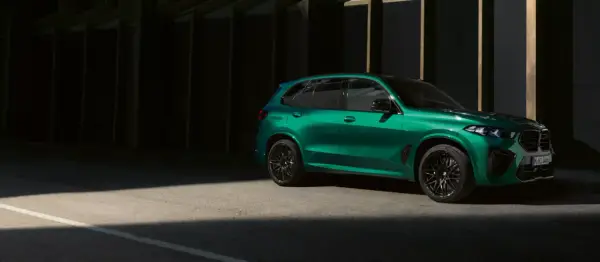 Zielony BMW X5 M z czarnymi felgami stoi na tle ciemnej architektury, oświetlony pasem światła.