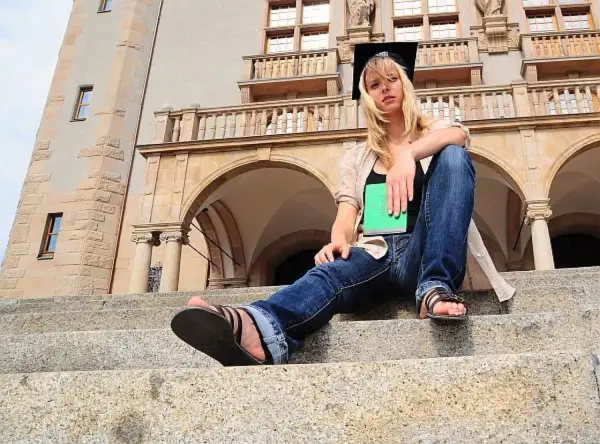 Studentka w czepku absolwenta z zieloną książką, siedząca na schodach przed budynkiem uniwersyteckim. Marzy o studiach zaocznych w Poznaniu.