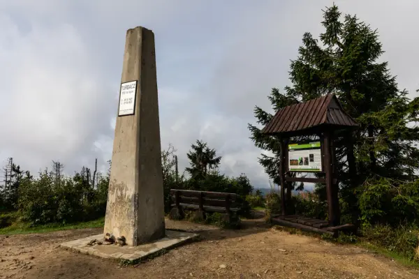 Kamienny obelisk na szczycie Turbacz w Gorcach, obok drewniana wiata z tablicą informacyjną i ławką.