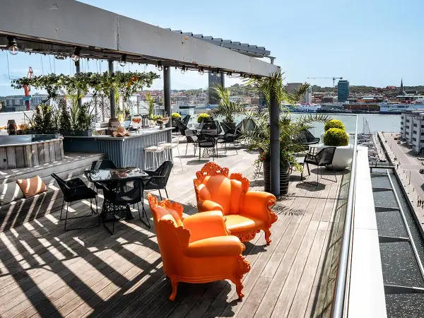 Solig rooftop bar i Göteborg med orangea fåtöljer och utsikt över hamnen.