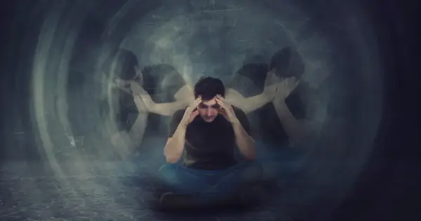 Mężczyzna siedzi ze zbolałą miną, trzymając się za głowę. Wokół niego unoszą się jego rozmazane, powielone sylwetki, symbolizujące schizofrenia objawy.