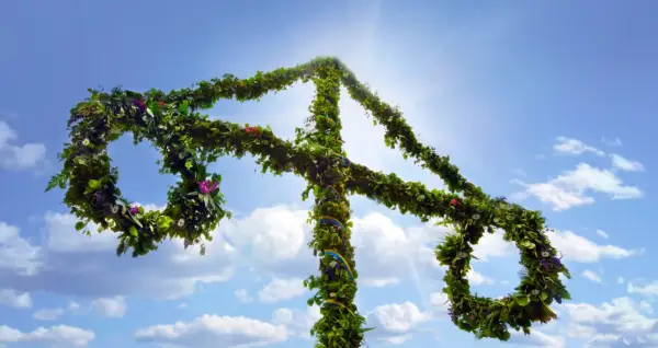 En midsommarstång, klädd i grönska och blommor, reser sig mot en klarblå himmel med solsken.