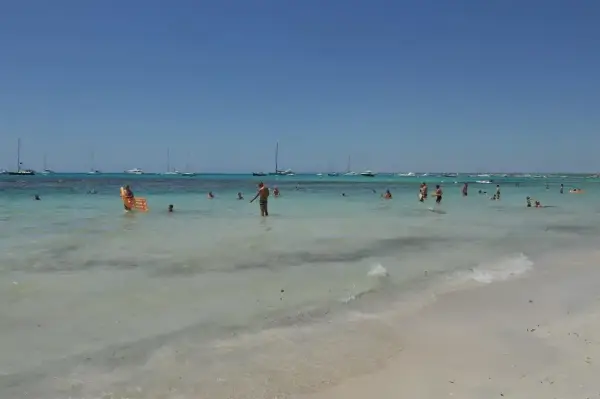 Słoneczny dzień na plaży Es Trenc. Ludzie kąpią się w turkusowej wodzie, a na horyzoncie widać żaglówki.