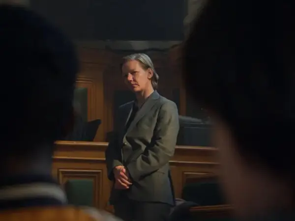 Sandra Hüller, con traje gris, se mantiene firme en una sala de audiencias, con las manos entrelazadas, mientras la cámara se enfoca en su rostro pensativo.