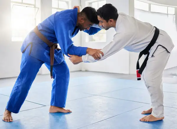 Dwóch mężczyzn w kimonach do BJJ, jeden w niebieskim z brązowym pasem, drugi w białym z czarnym pasem, przygotowuje się do walki.