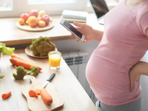 Ciężarna kobieta zmagająca się z brakiem apetytu w ciąży, szuka w telefonie przepisu na zdrowe jedzenie. Na blacie warzywa i sok.