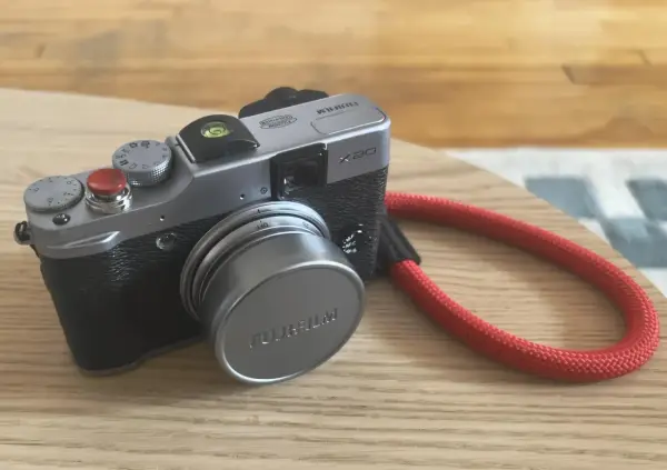 Srebrny aparat Fujifilm X20 z czerwoną smyczą leży na drewnianym blacie.