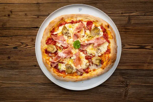 Eine köstliche Pizza mit Prosciutto Crudo, Artischocken und frischem Basilikum auf einem weißen Teller, serviert auf rustikalem Holz.