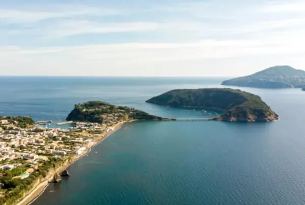 Panoramica di un'isoletta tra Napoli e Ischia, con un borgo costiero, un porto turistico e un ponte che collega a un'altra isola verdeggiante.
