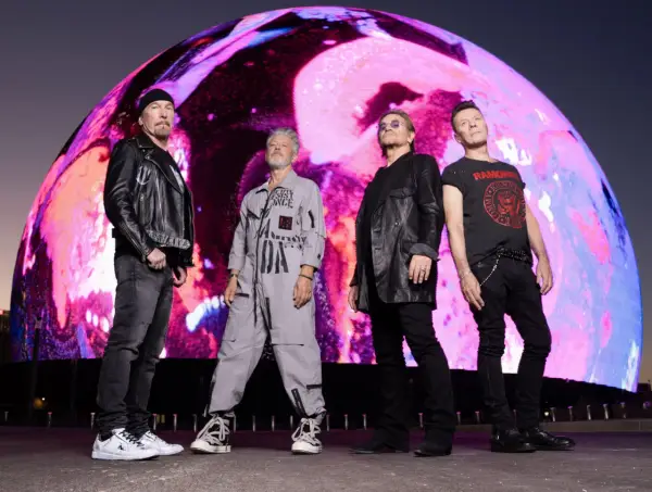 U2 w nowej płycie. Czterech członków zespołu stoi przed gigantycznym, kolorowym ekranem.