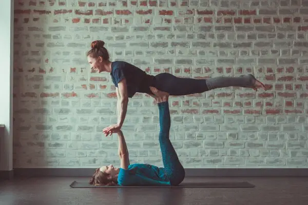 Dwie kobiety ćwiczą **acroyoga** na macie. Jedna leży na plecach, podtrzymując drugą, która unosi nogi w powietrzu.