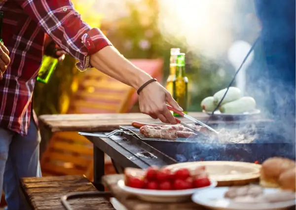 Hombre asa salchichas envueltas en tocino, ideal para saber qué llevar a una barbacoa para sorprender. Hay cervezas y tomates en la mesa.