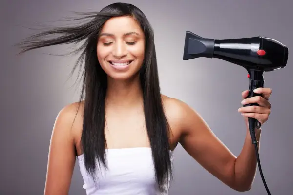 Mujer sonríe mientras se seca el pelo fino y escaso con un secador negro.