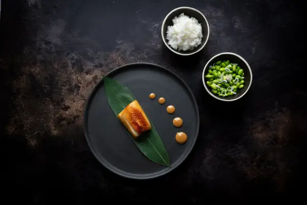 A perfectly cooked nobu miso black cod rests on a dark plate with a green leaf. Drizzled sauce and sides of rice and edamame complete the elegant dish.
