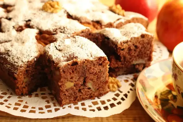 Kawałki aromatycznego ciasta cynamonowego z orzechami, posypane cukrem pudrem, obok jabłek i filiżanki herbaty.