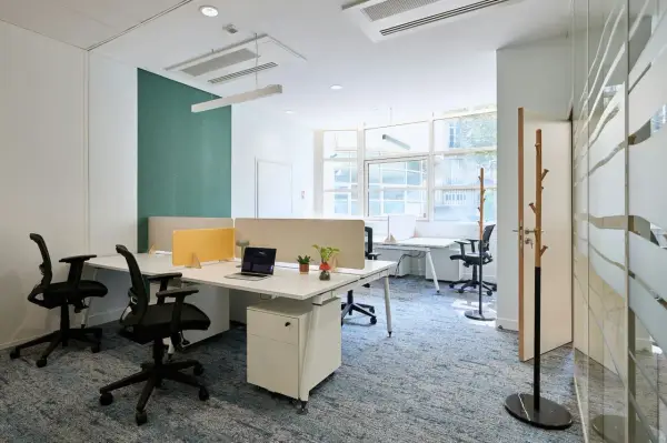 Un bureau chaleureux avec des postes de travail modernes, des cloisons acoustiques et un tapis bleu doux. L'espace est lumineux grâce aux grandes fenêtres.