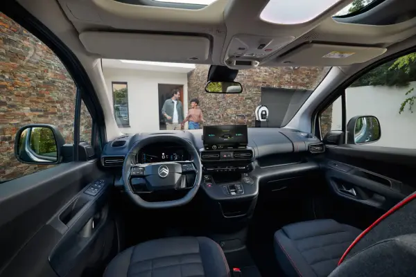 Interior de la nueva Citroën Berlingo, con pantalla táctil, volante moderno y asientos cómodos. Afuera, una pareja se acerca a un punto de carga.