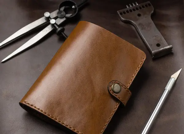Brązowy portfel ze skóry, narzędzia do obróbki skóry: cyrkiel, dziurkacz, nożyk. Leather cuir to znaczy skóra.