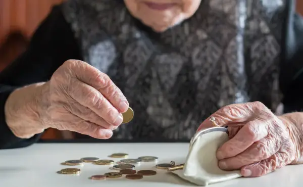 Dłoń starszej kobiety z pierścionkiem wkłada monetę do portmonetki. Rozrzucone monety na stole sugerują trudności finansowe, być może związane z emeryturą po mężu.