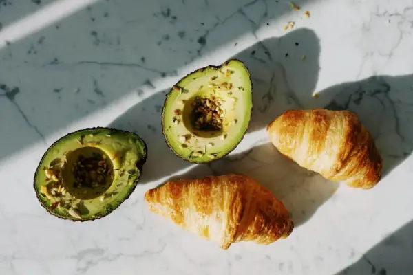 Dwa połówki awokado z nasionami i dwa chrupiące croissanty na marmurowym blacie. Idealne składniki na pyszne awokado przepisy.
