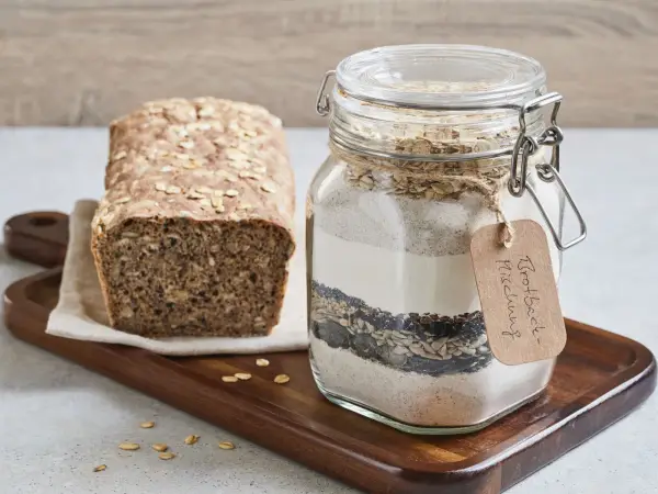 Selbstgemachte brotbackmischung im Glas, bereit zum Backen. Daneben ein frisch gebackenes Brot mit Haferflocken und Körnern.