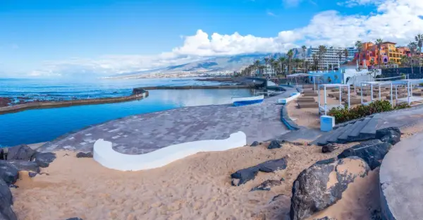 Słoneczna plaża w Playa de las Américas z basenem, łódką i kolorowymi budynkami w tle.
