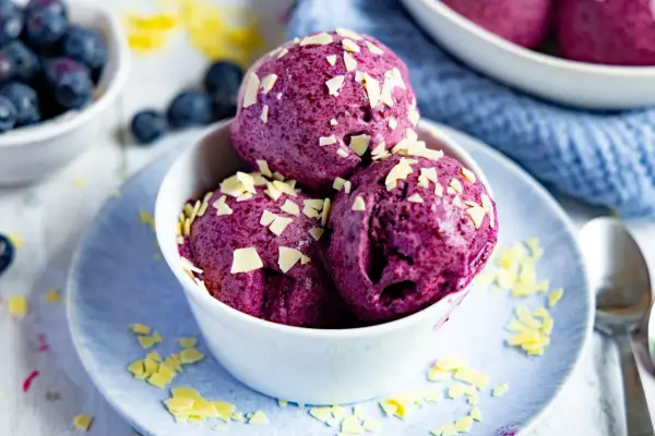Drei Kugeln Blaubeer Eis, garniert mit weißen Schokostückchen, in einer weißen Schale auf einem blauen Teller.