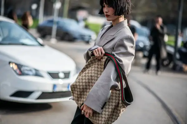 Kobieta w szarym żakiecie trzyma torbę z monogramem Gucci, jakby to był certyfikat autentyczności.