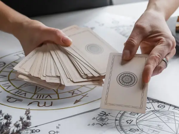 Dłonie trzymają talię kart, przygotowując się do wróżenia. Rozłożone na stole diagramy astrologiczne sugerują, jak stawiać tarota.