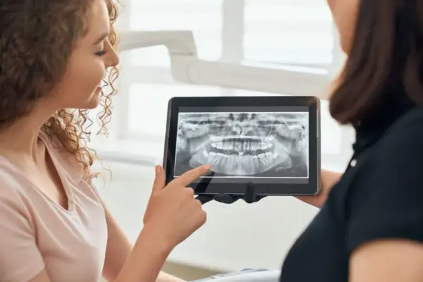 Pacjentka z kręconymi włosami ogląda z dentystką panoramiczny rentgen stomatologiczny na tablecie.