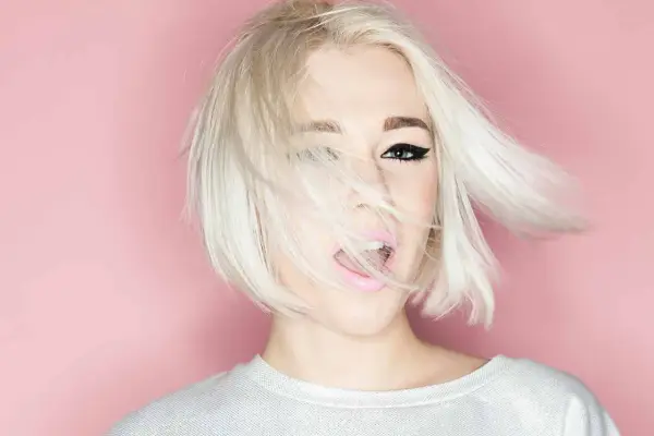 Mujer con cabello rubio platino y corte recto al hombro, con un ojo visible y labios rosados abiertos.