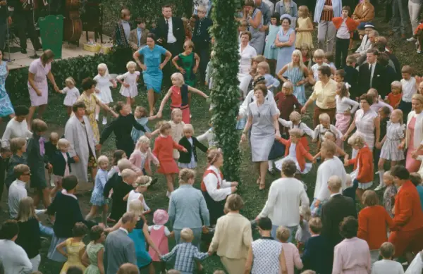 Många barn och vuxna dansar ringdans runt en midsommarstång i Sverige.