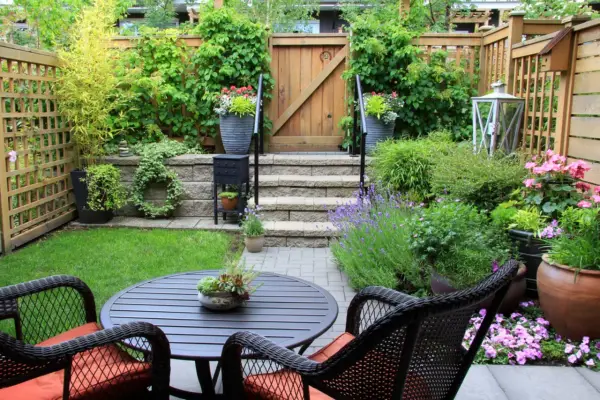 Ein kleiner Garten mit Sitzgruppe, Stufen und Holztor. Üppige Pflanzen und Blumen schaffen eine grüne Oase.