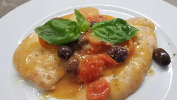 Delizioso pollo alla mediterranea con pomodorini, olive nere e basilico fresco su un piatto bianco.