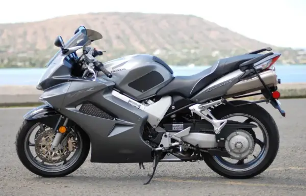 Szary motocykl Honda VFR 800 z widokiem na wodę i wzgórza w tle.