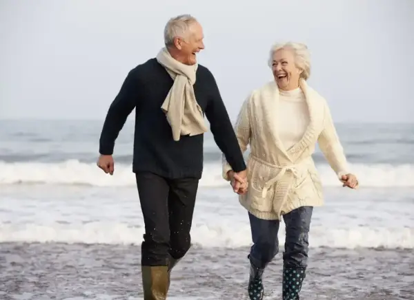Szczęśliwa para seniorów biegnie plażą, trzymając się za ręce. Idealne tanie wycieczki dla seniorów, by poczuć radość życia!