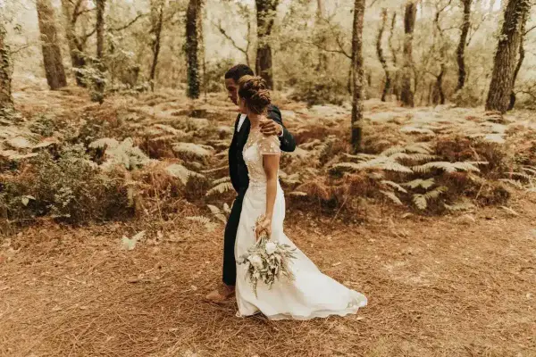 Un couple se promène dans une forêt, la mariée porte une robe blanche et tient un bouquet. Une scène de formalité mariage en pleine nature.